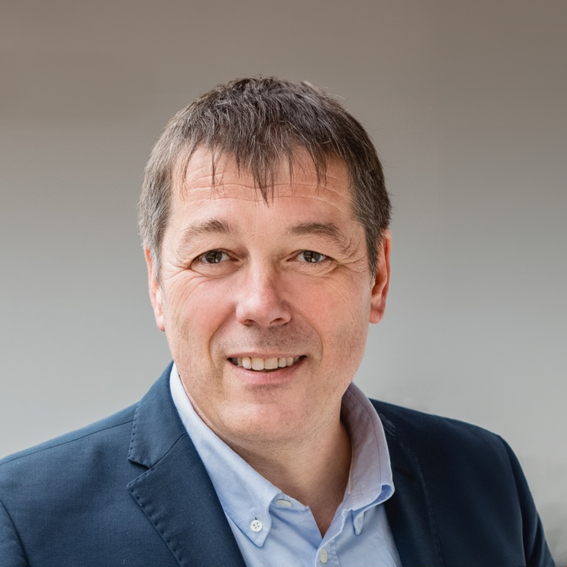 Porträt eines lächelnden Mannes im Anzug mit Hemd vor neutralem Hintergrund.