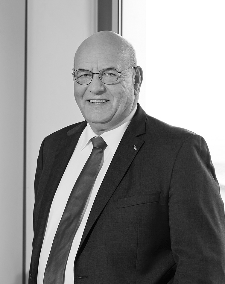 Porträt eines älteren Mannes mit Brille im Anzug und Krawatte, lächelnd vor hellem Hintergrund.