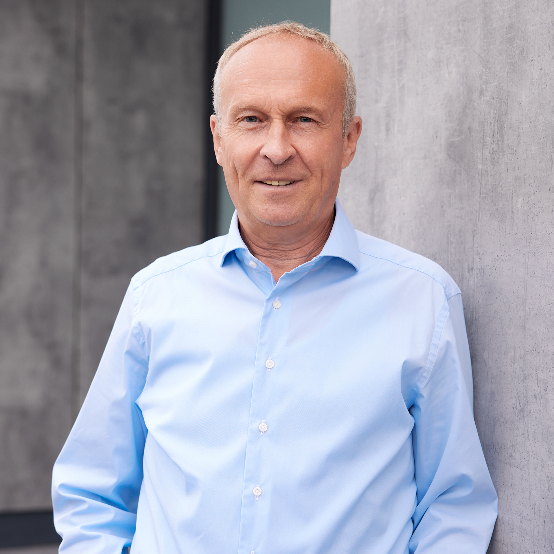 Porträt eines älteren Mannes im hellblauen Hemd, stehend vor einer Betonwand.
