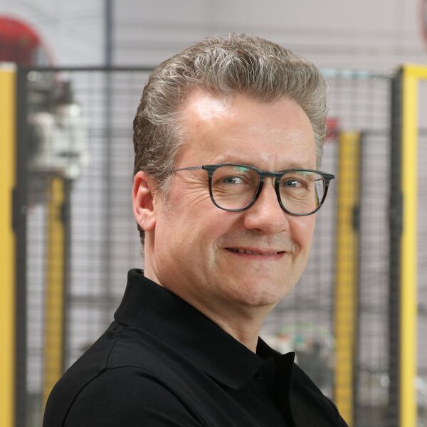 Porträt eines lächelnden Mitarbeiters mit Brille und schwarzem Poloshirt in einer Produktionsumgebung.