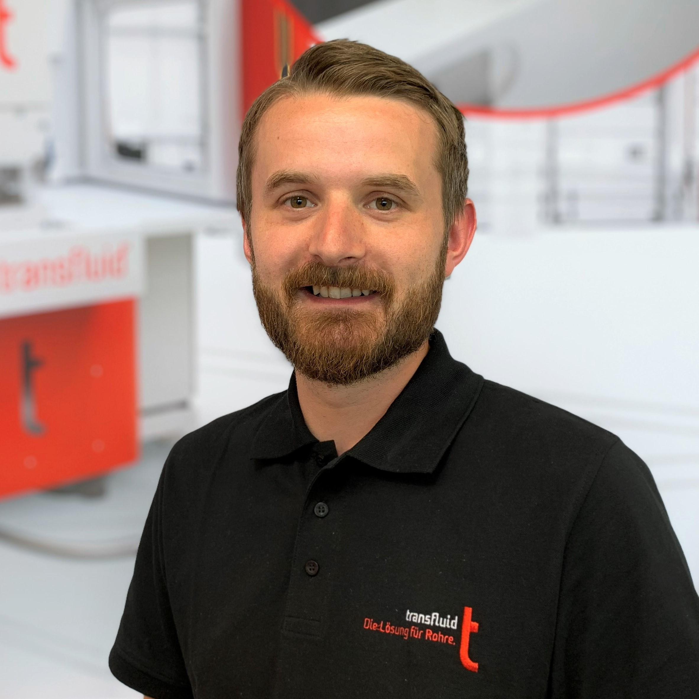 Porträt eines Mitarbeiters mit Bart und schwarzem Poloshirt vor einer Maschine.