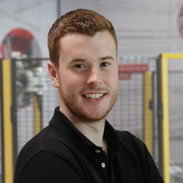 Mitarbeiter mit kurzem Haar und Bart im schwarzen Poloshirt vor Industrieanlage.