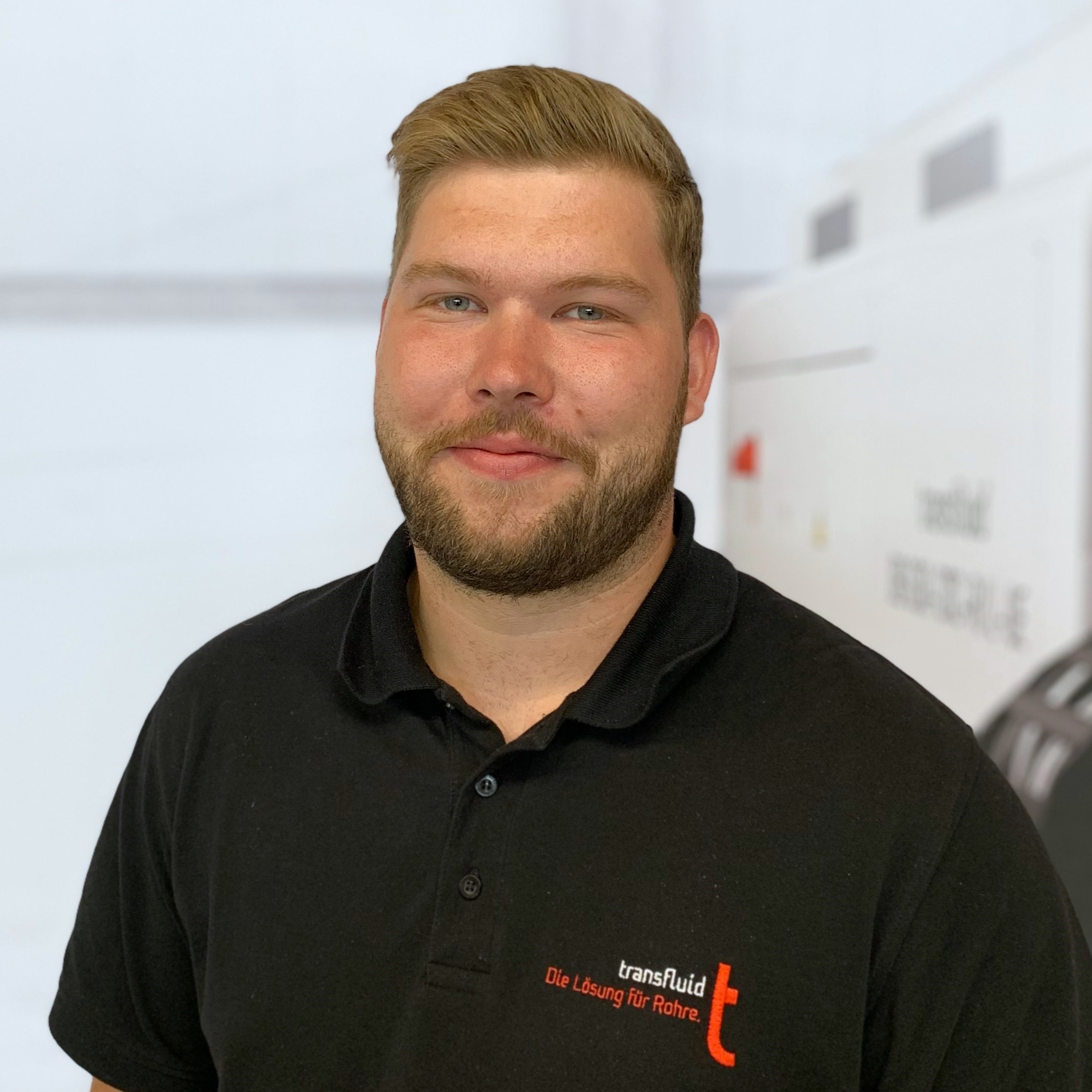 Mitarbeiter mit kurzem Haar und Bart im schwarzen Poloshirt in heller Industrieumgebung.