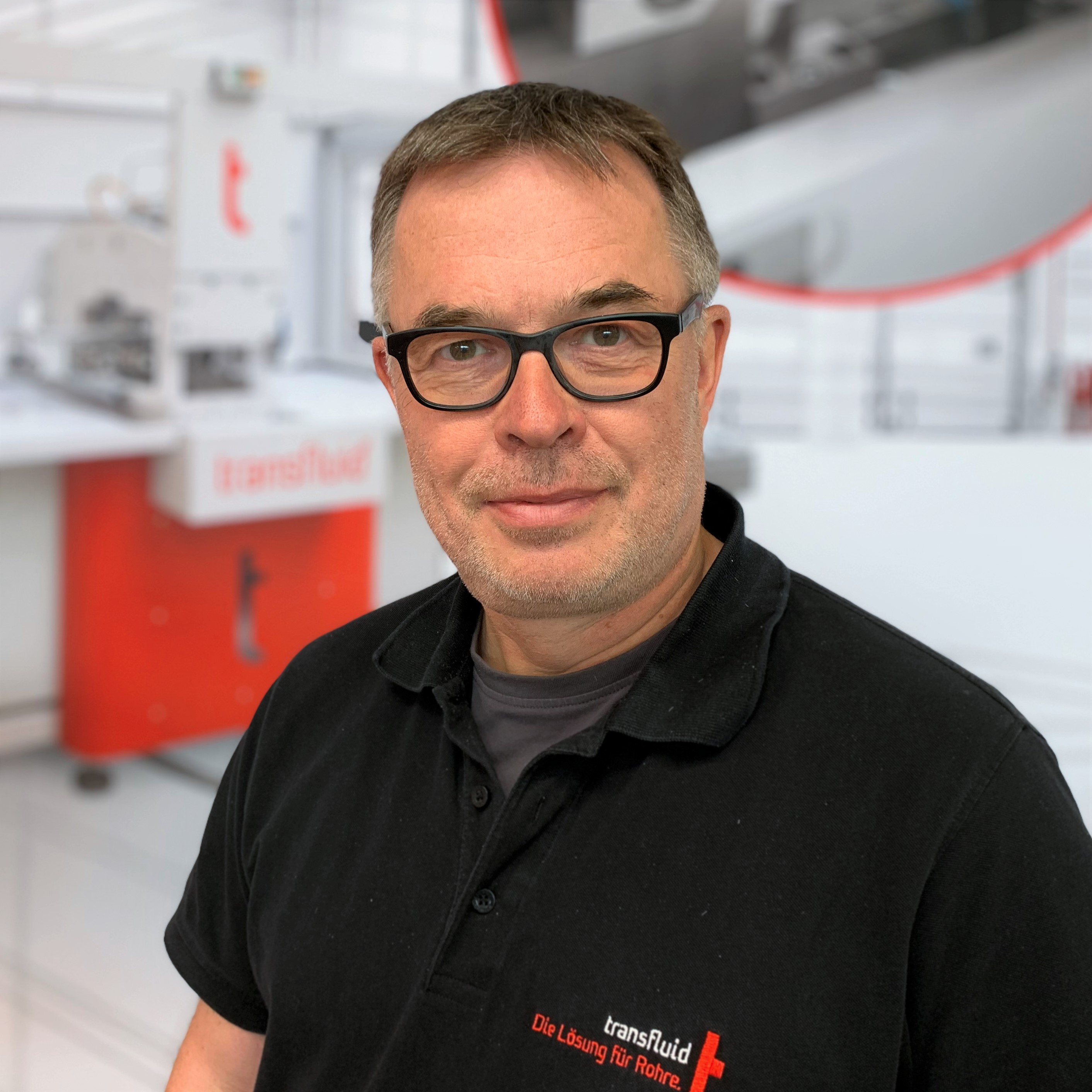 Mitarbeiter mit Brille und schwarzem Poloshirt vor unscharfer Industrieanlage.