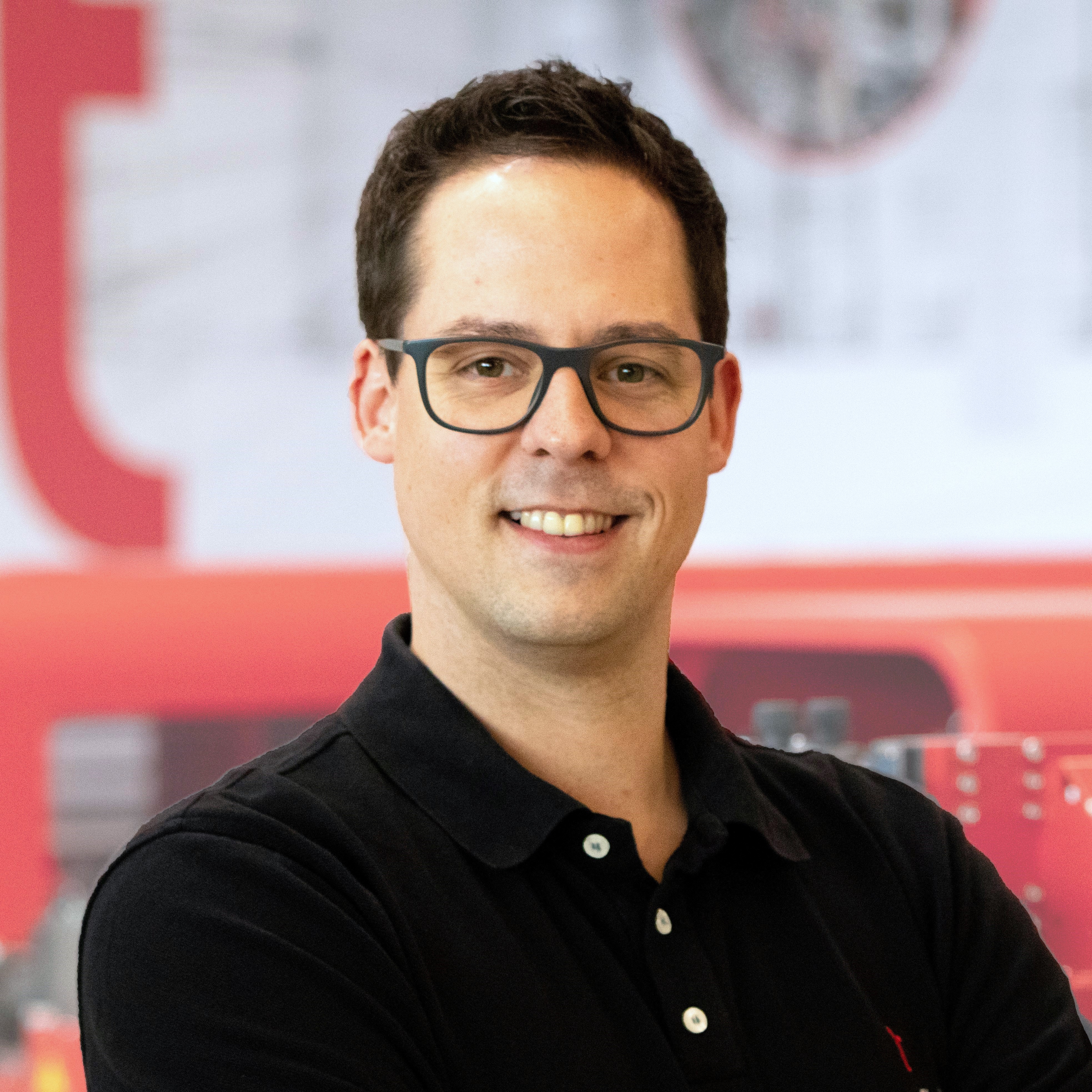Mitarbeiter mit Brille und schwarzem Poloshirt vor roter Industrieanlage.
