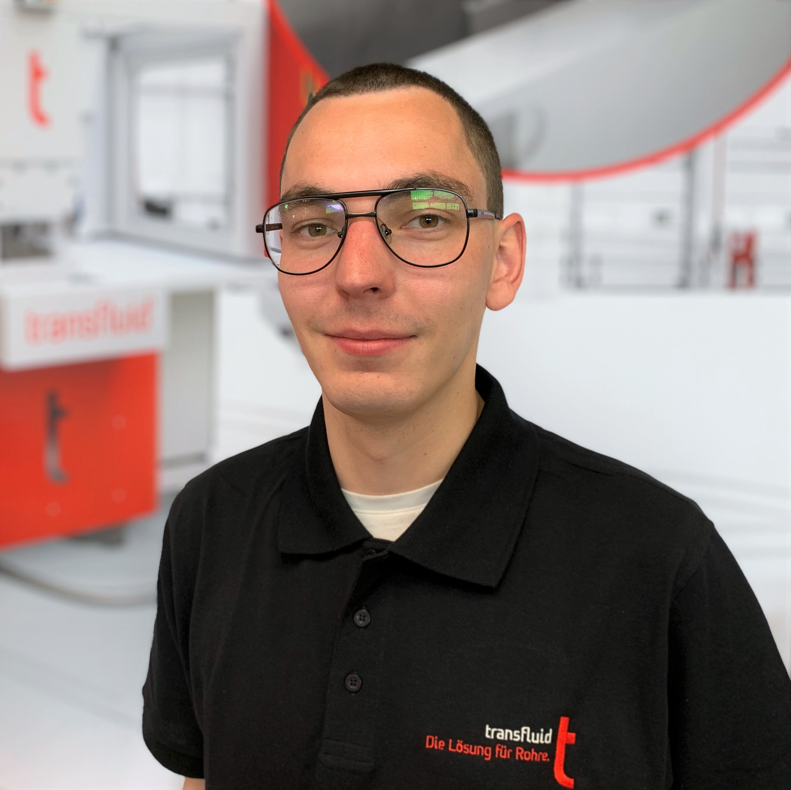 Mitarbeiter mit Brille und schwarzem Poloshirt vor unscharfer Industrieanlage.