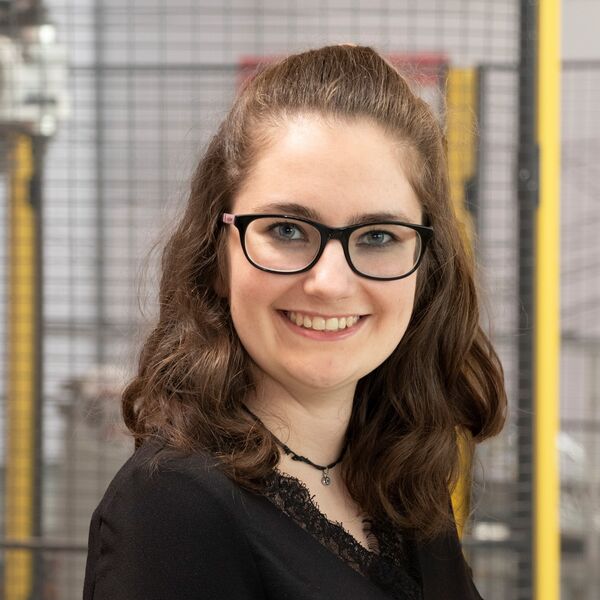 Mitarbeiterin mit Brille und dunklem Oberteil vor unscharfer Industrieanlage.