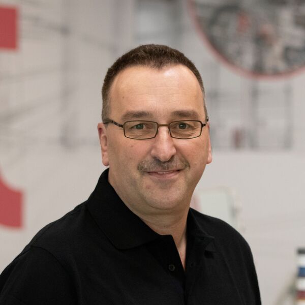 Mitarbeiter mit Brille und schwarzem Poloshirt vor unscharfer Industrieanlage.