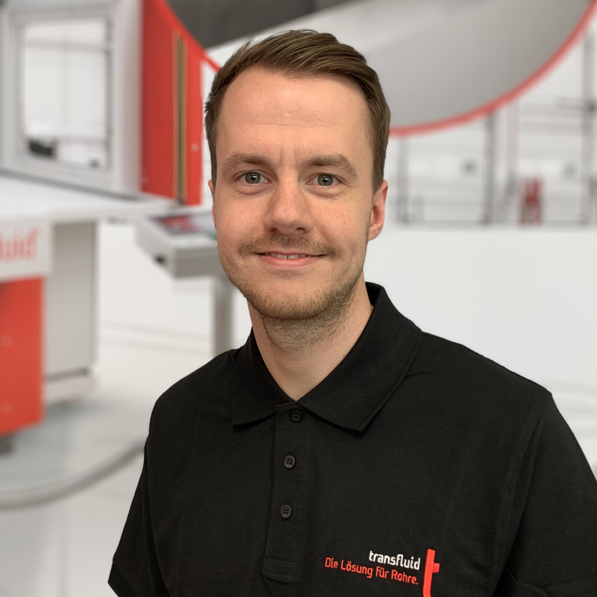 Mitarbeiter im schwarzen Poloshirt vor unscharfer Industrieanlage.