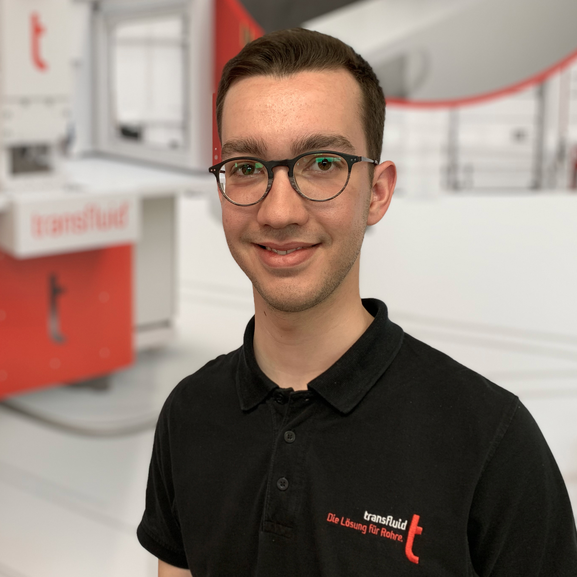 Mitarbeiter mit Brille im schwarzen Poloshirt vor unscharfer Industrieanlage.