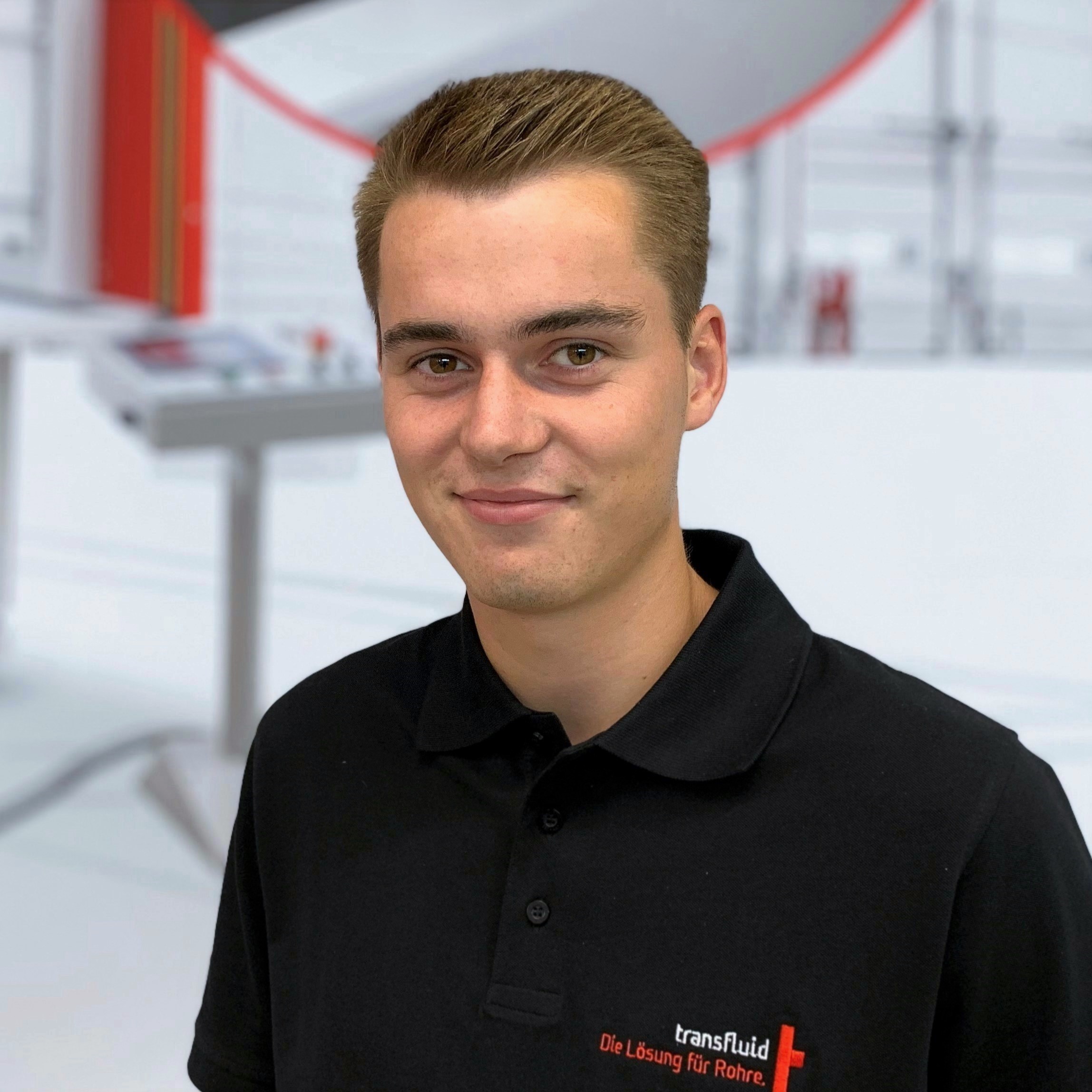 Mitarbeiter im schwarzen Poloshirt vor unscharfer Industrieanlage.