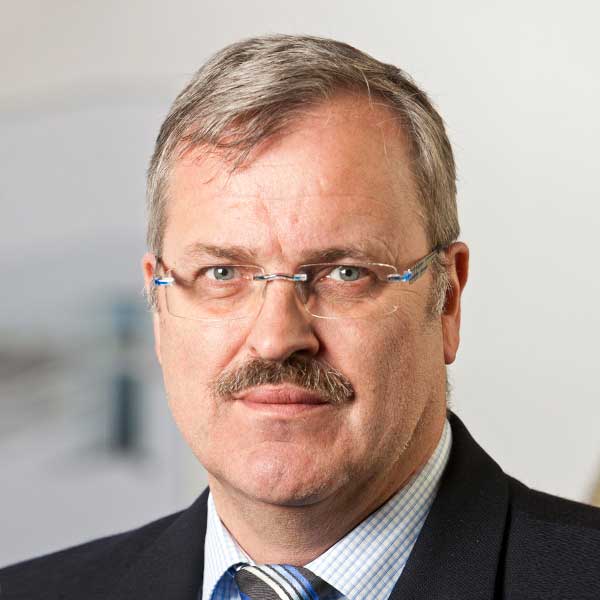 Mitarbeiter mit Brille, Schnurrbart und Anzug vor hellem Hintergrund.