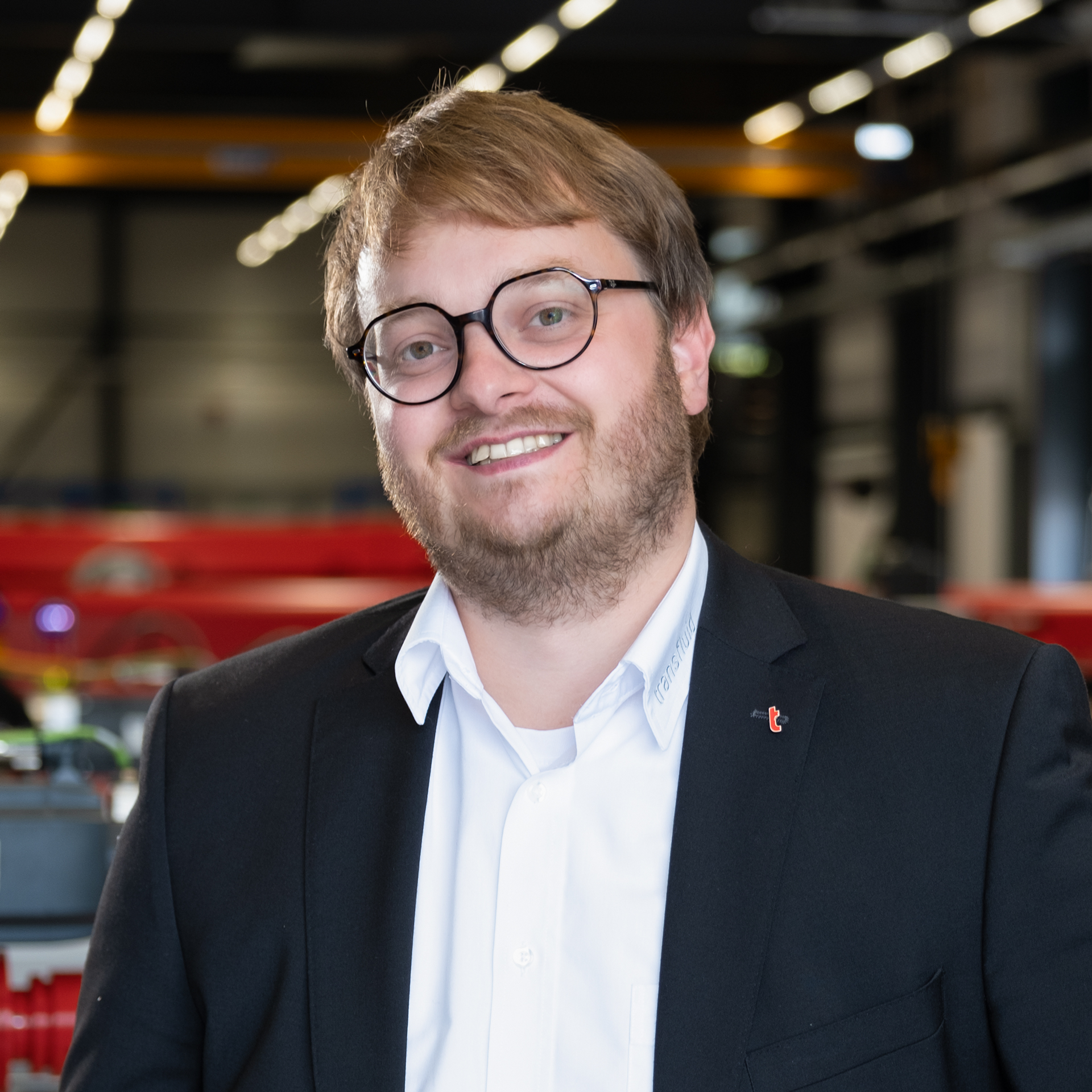 Mitarbeiter mit Brille im dunklen Sakko in moderner Industriehalle mit unscharfem Hintergrund.
