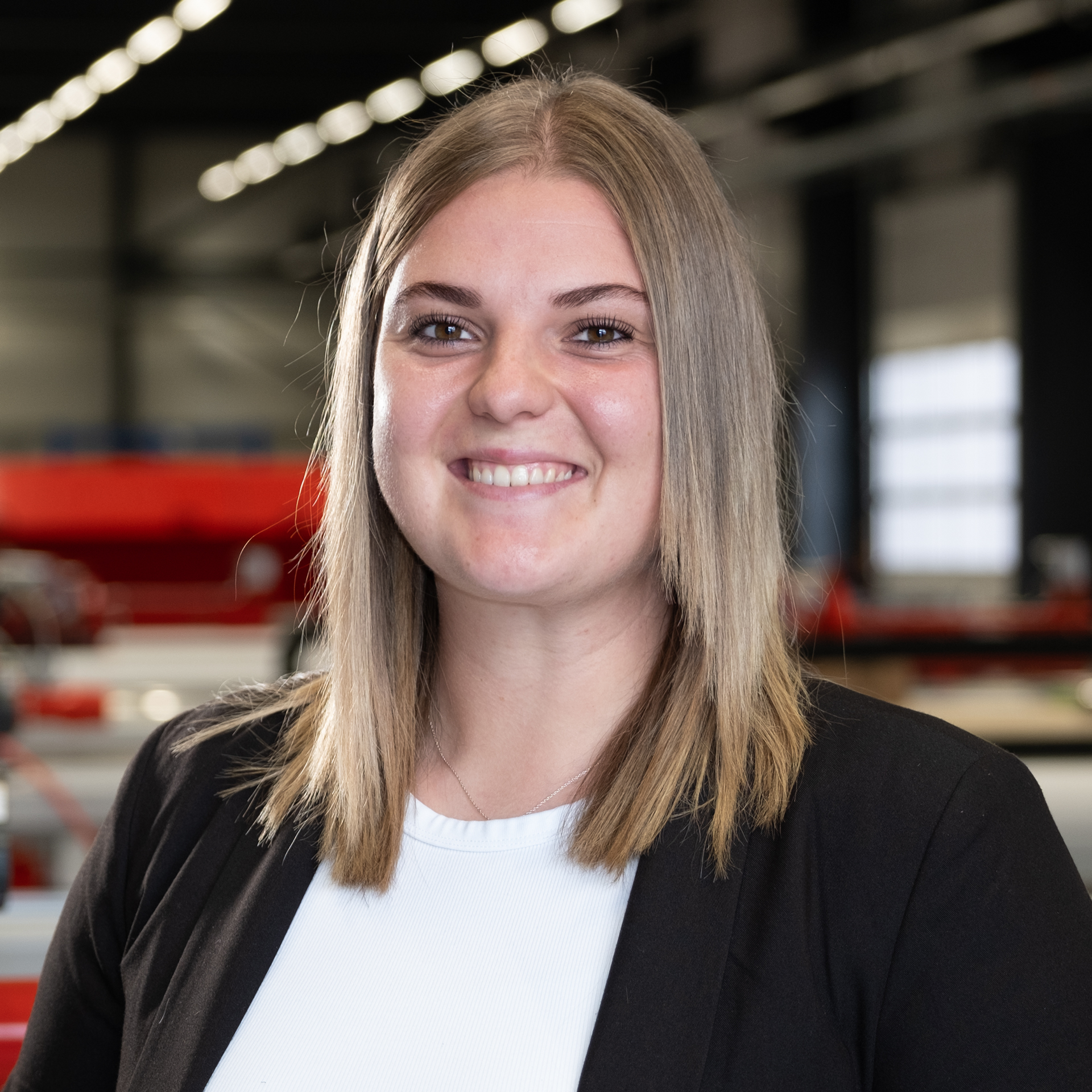 Junge Mitarbeiterin im Blazer lächelt in Industriehalle mit Maschinen im Hintergrund.